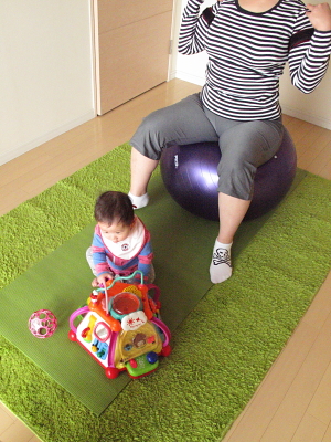 バランスボールでトレーニングする隣で、赤ちゃんがおもちゃで遊んでいる様子